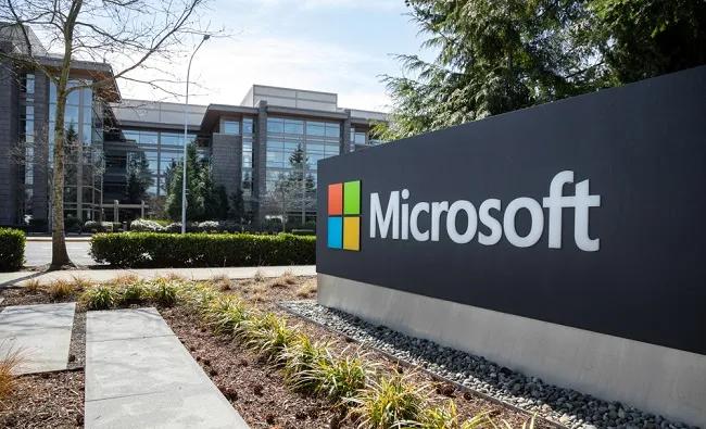 Microsoft logo sign in front of modern campus headquarters building, featuring colorful windowed panes and green landscaping.