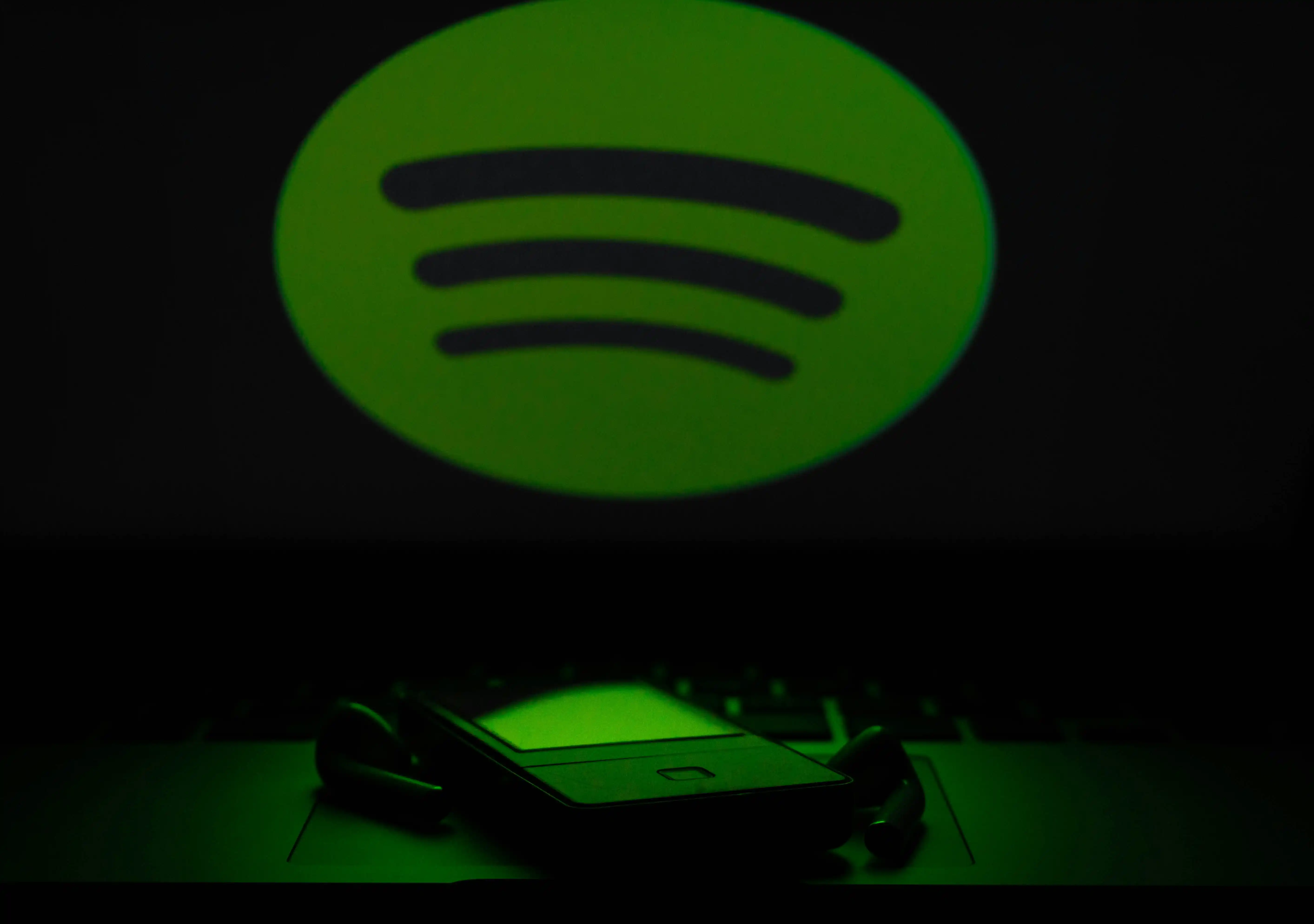 Spotify logo glowing bright green in dark background with smartphone and wireless earbuds resting on laptop keyboard.