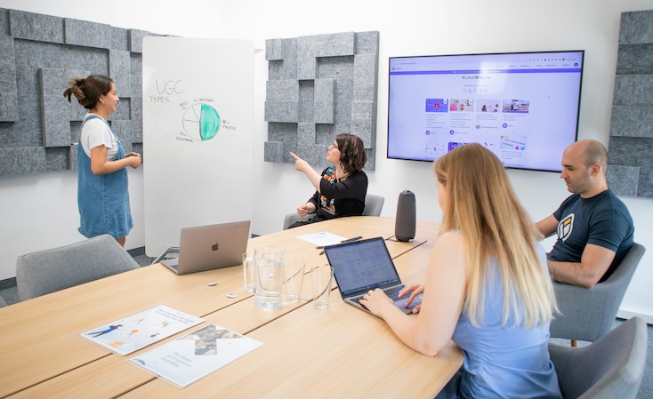 A software development team collaborates in a room with a whiteboard and a TV