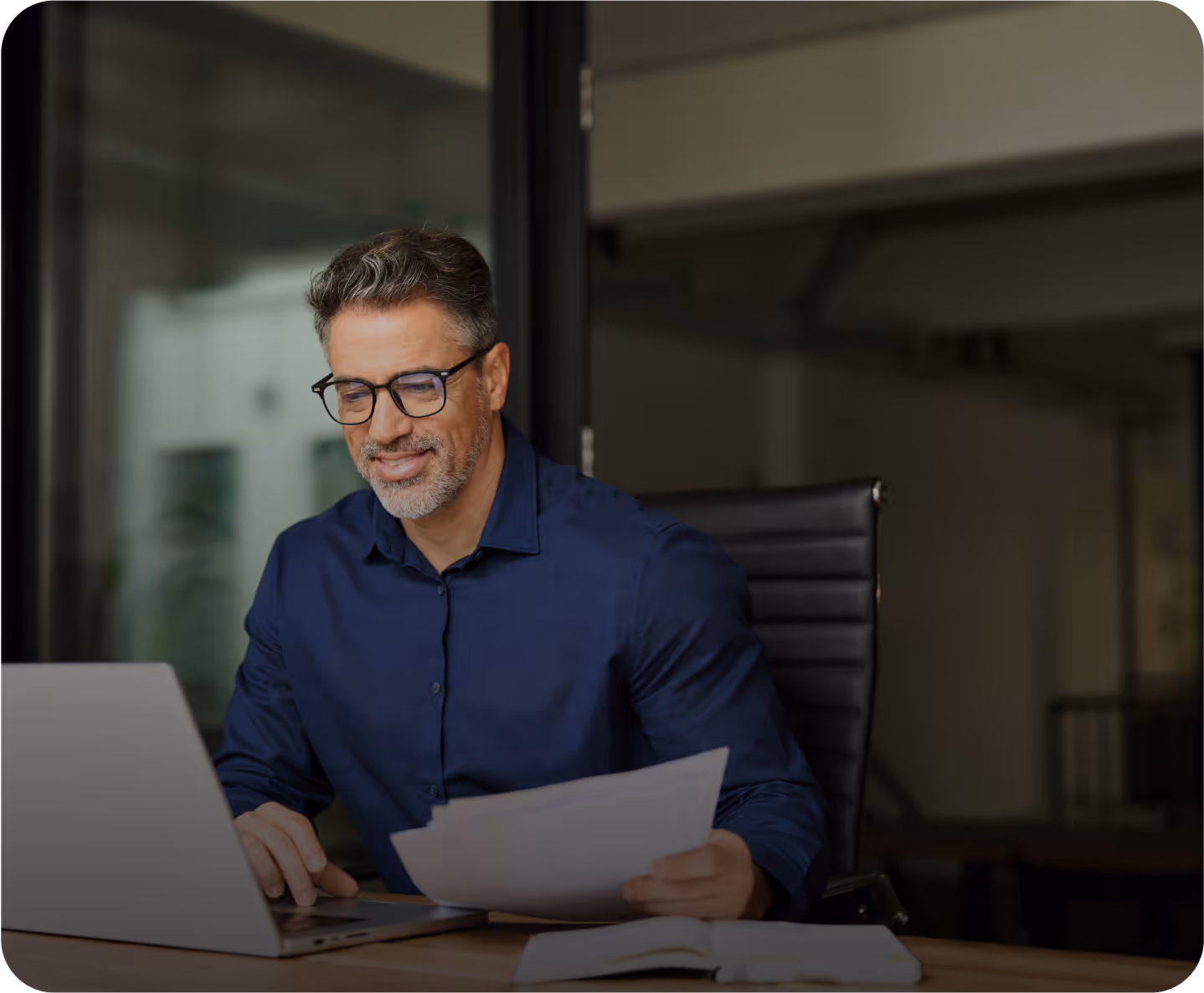 A man in glasses and a blue shirt works on a laptop while holding documents.
