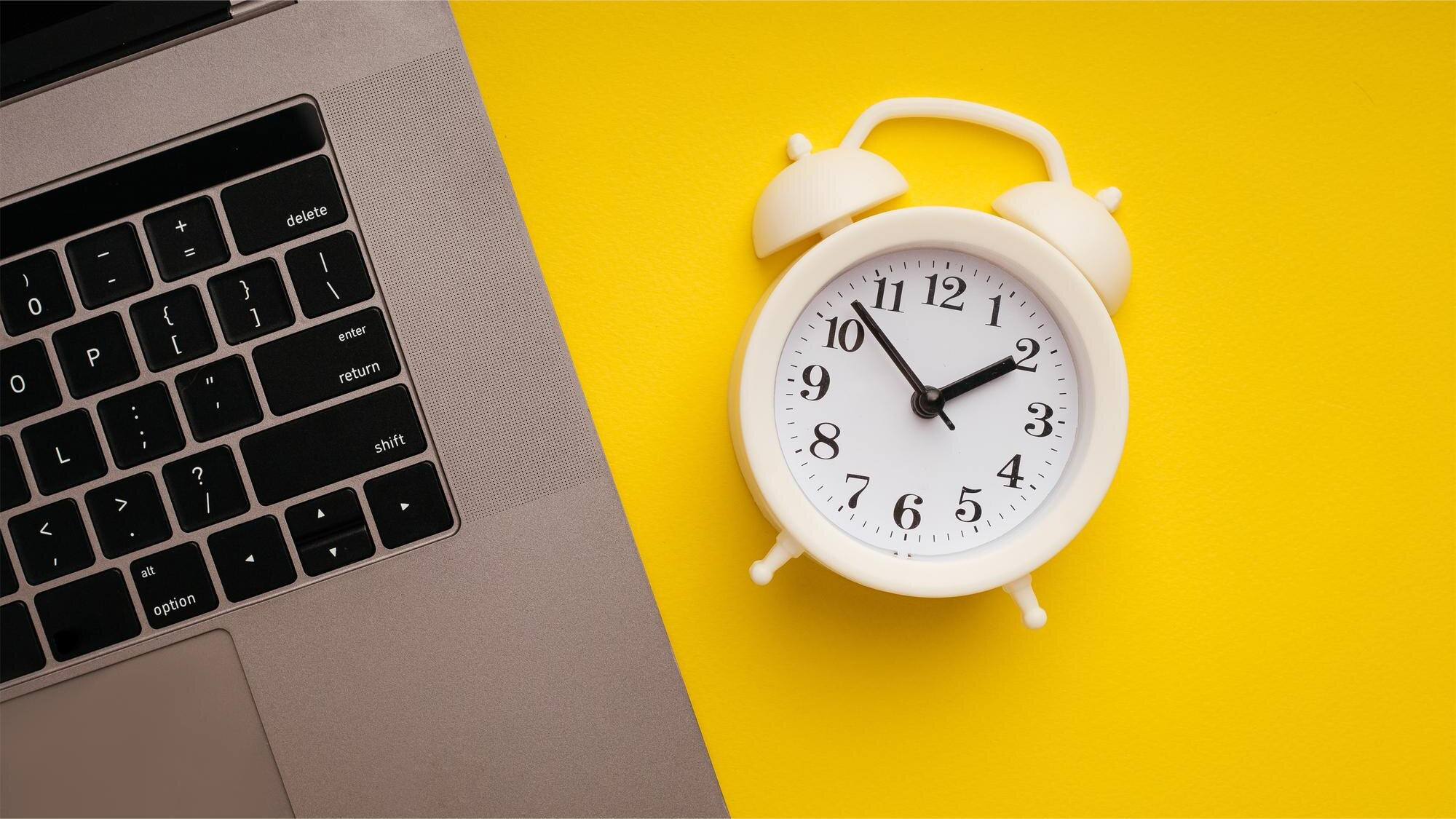 White alarm clock and laptop on yellow background – symbolizing the ISO 27001:2022 vs 2013 transition deadline countdown