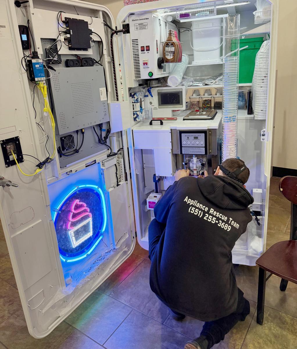 Technician wearing black hoodie labeled 'Appliance Rescue Team' kneeling and working inside an open frozen yogurt or ice cream machine with neon-lit soft serve icon.