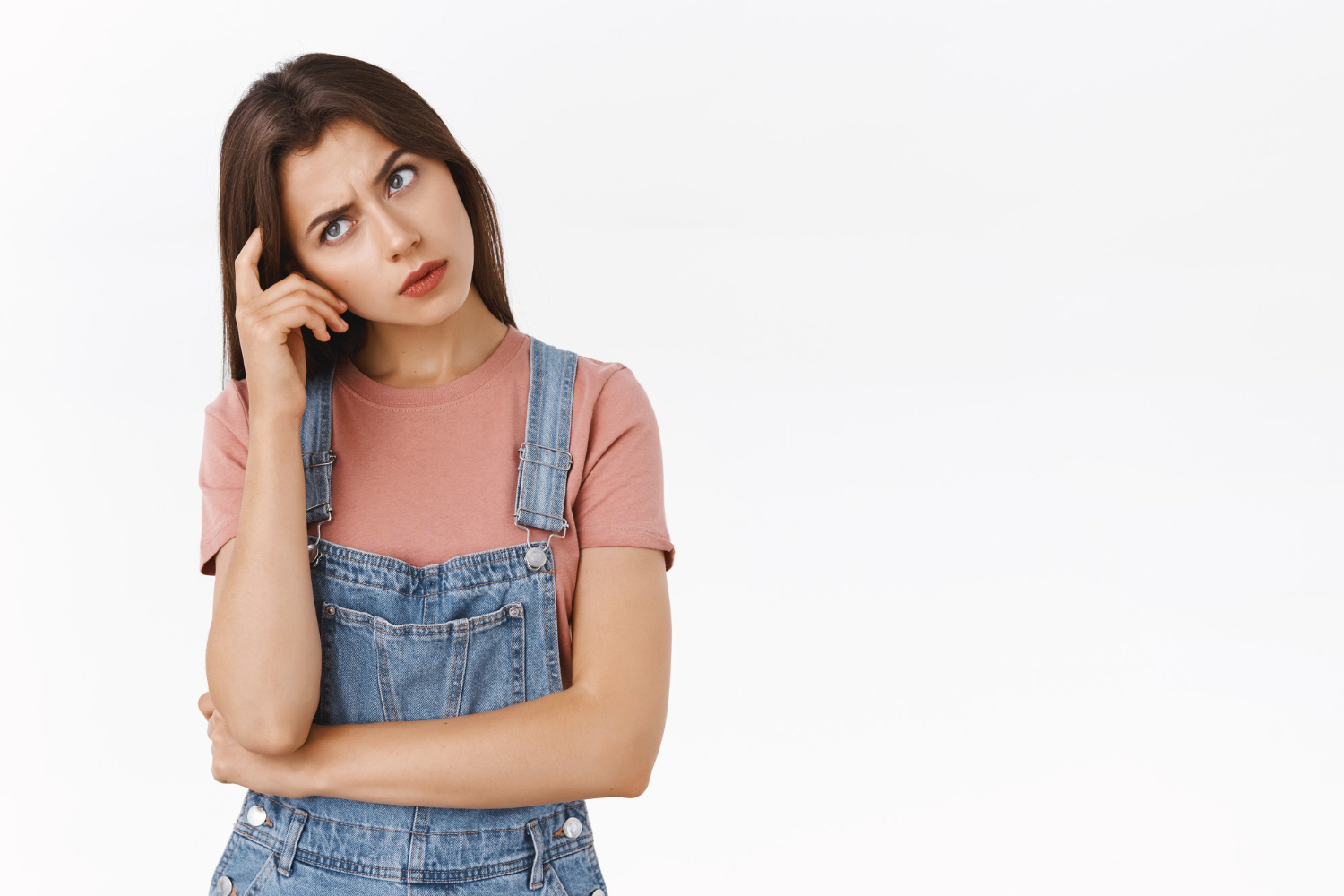 Confused young woman in a pink t-shirt and denim overalls scratching her head and frowning, looking thoughtful while trying to make a decision.