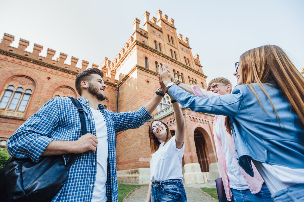 Group of happy college students standing outside a historic university building, smiling and giving each other high-fives in celebration