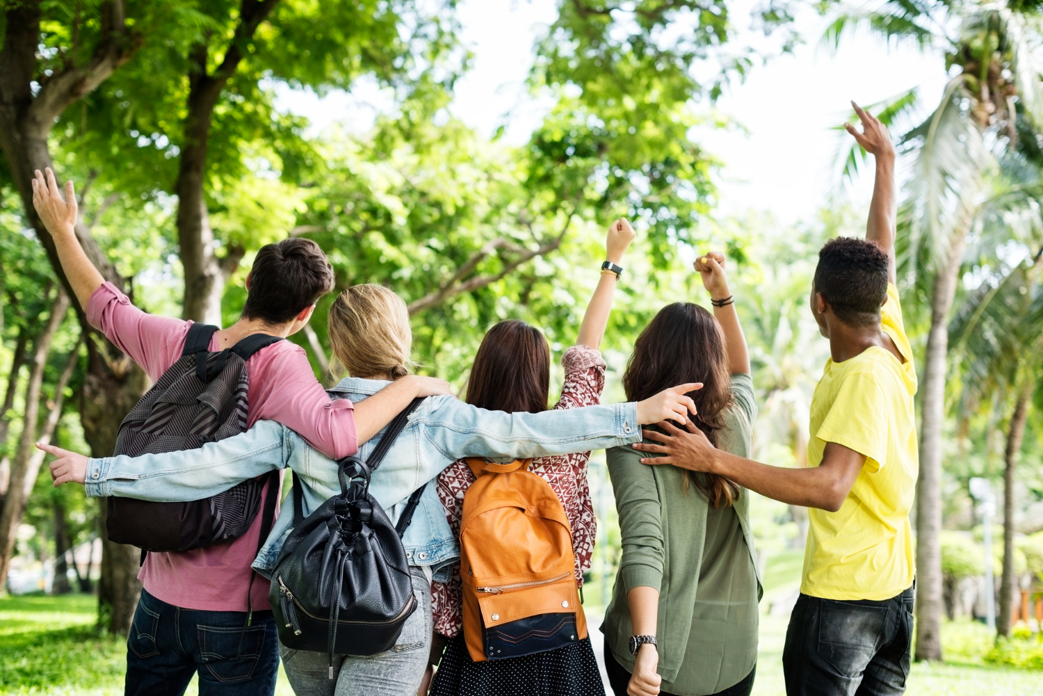 Group of diverse students with backpacks standing outdoors in a park, arms around each other, cheering and raising their hands in celebration.