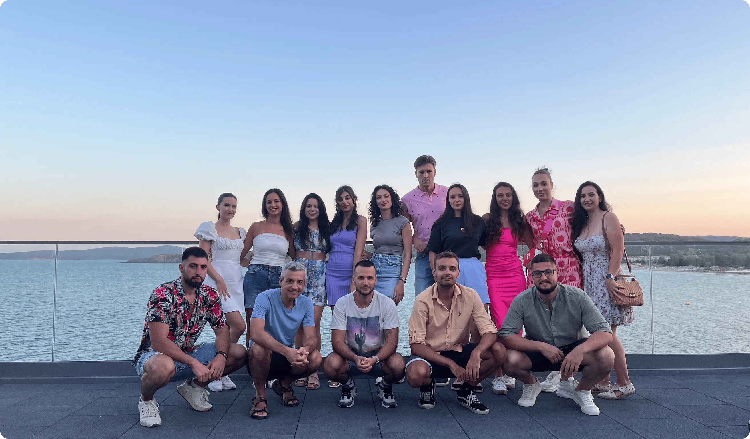 Group of fifteen young adults posing on a terrace overlooking a calm sea at sunset.