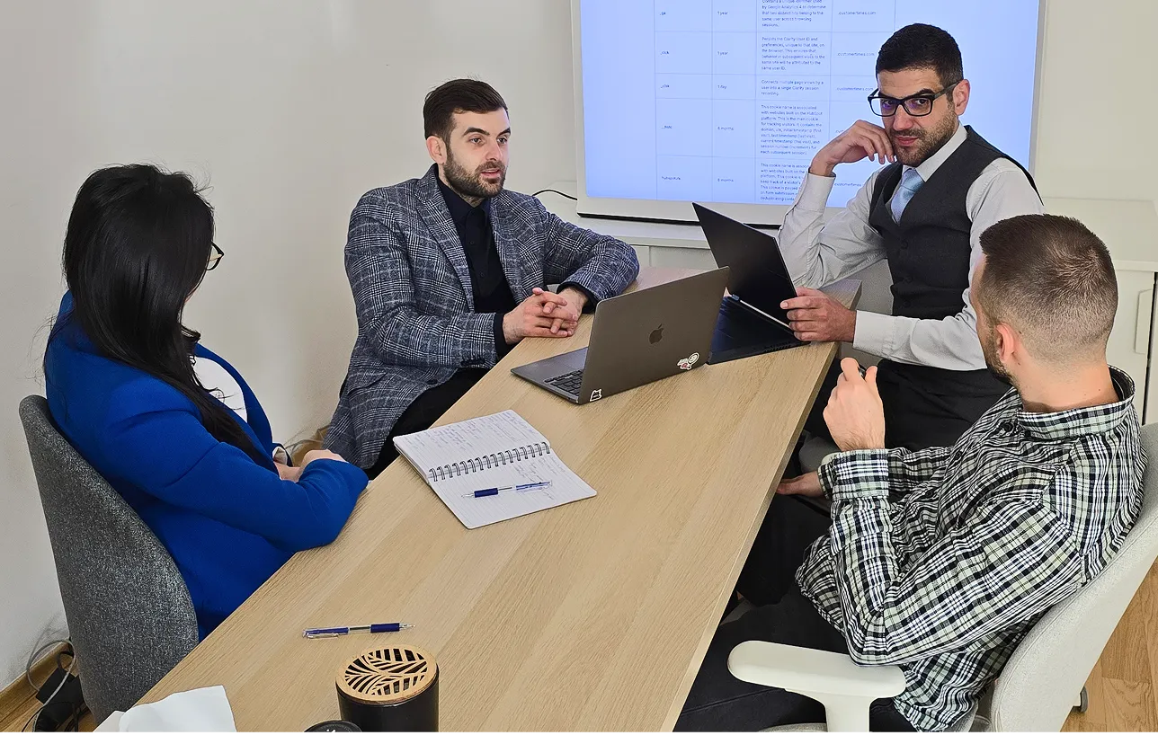 Four professionals sitting around a conference table in a meeting, two using laptops and a large screen displaying data in the background.