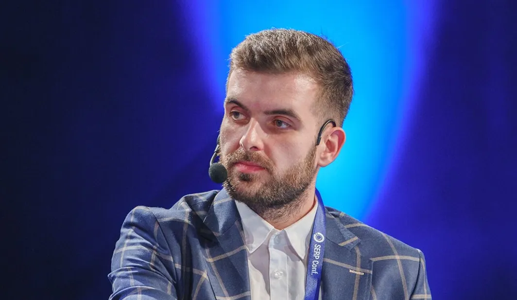 Man with a beard wearing a blue checkered suit jacket, white shirt, and headset microphone against a blue-lit background.