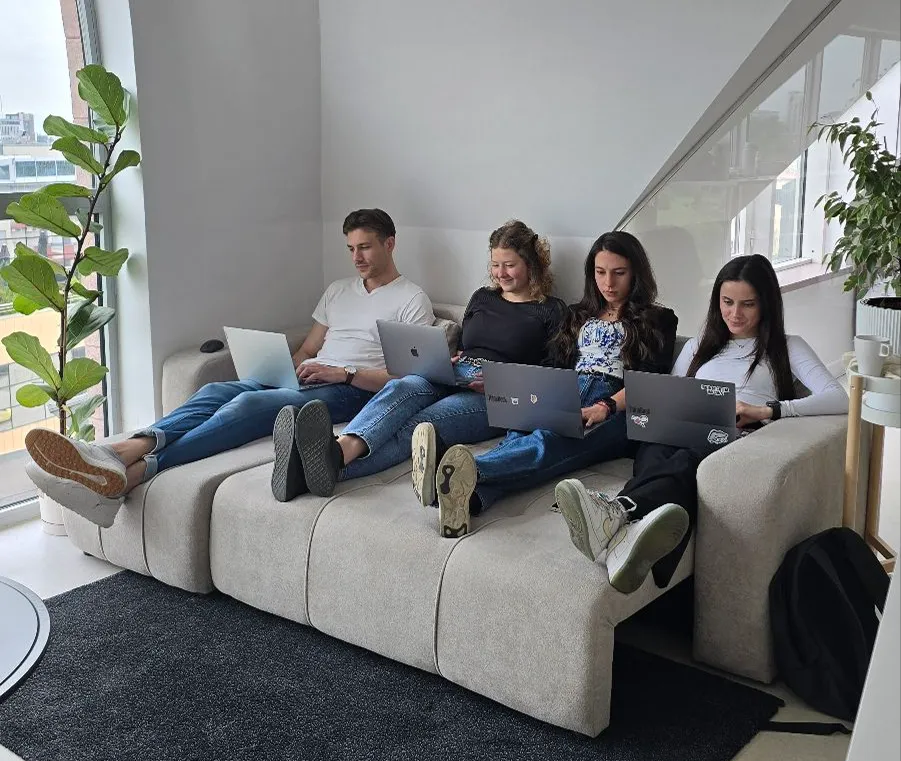 Four young adults sitting on a beige couch with laptops, working together in a bright modern room with plants nearby.