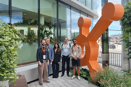 Five people standing outside a building next to a large orange sculpture and greenery.