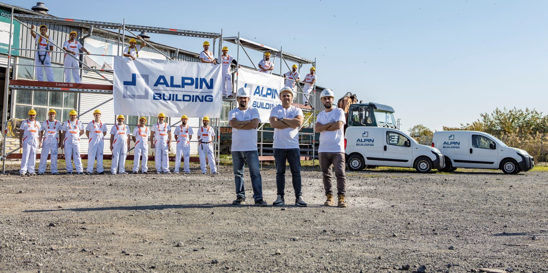 Construction team of Alpin Building with vehicles and scaffolding – Group photo of construction workers and engineers wearing branded uniforms and safety gear, posing in front of a construction site with scaffolding, company banners, and branded vans. Keyw