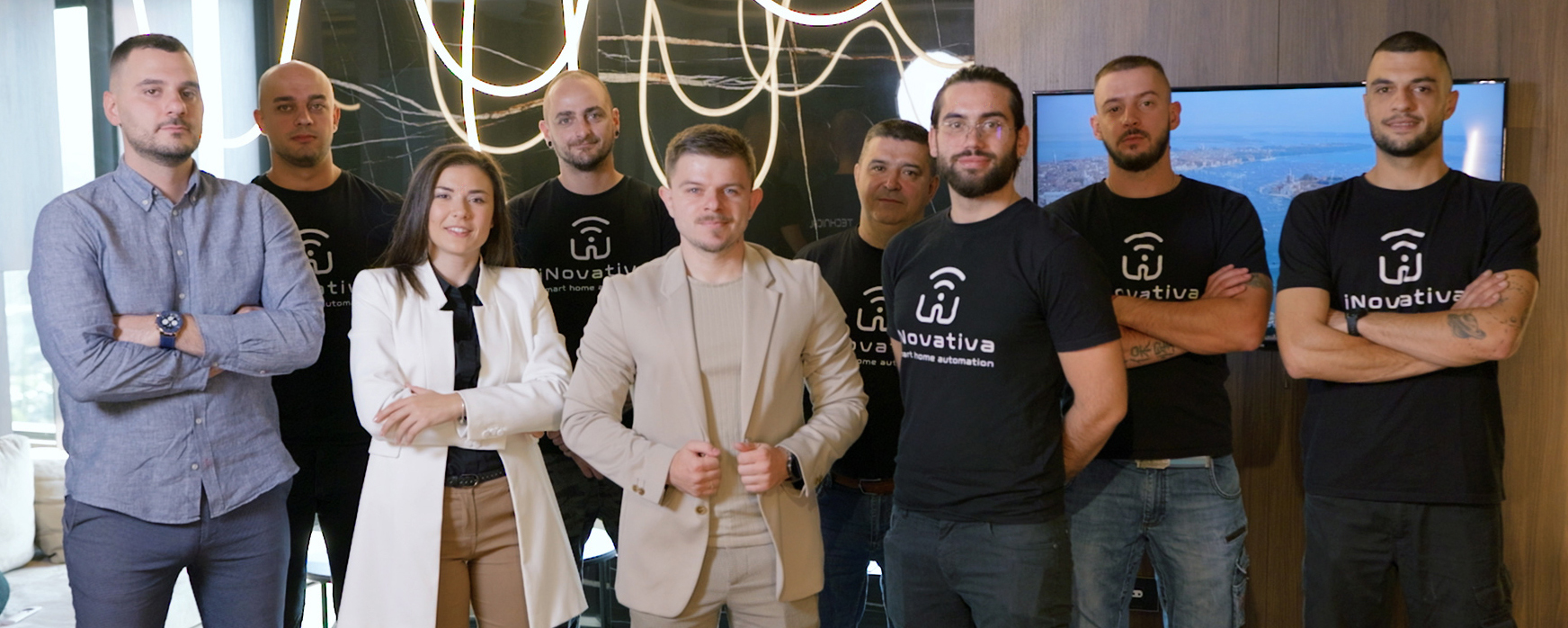 Innovativa smart home automation team in modern office setting – Team photo of eight professionals, some in branded Innovativa t-shirts, standing confidently in a stylish office with modern lighting and a TV screen displaying a cityscape. Keywords: smart home automation team, Innovativa, startup team, office portrait, modern workspace.
