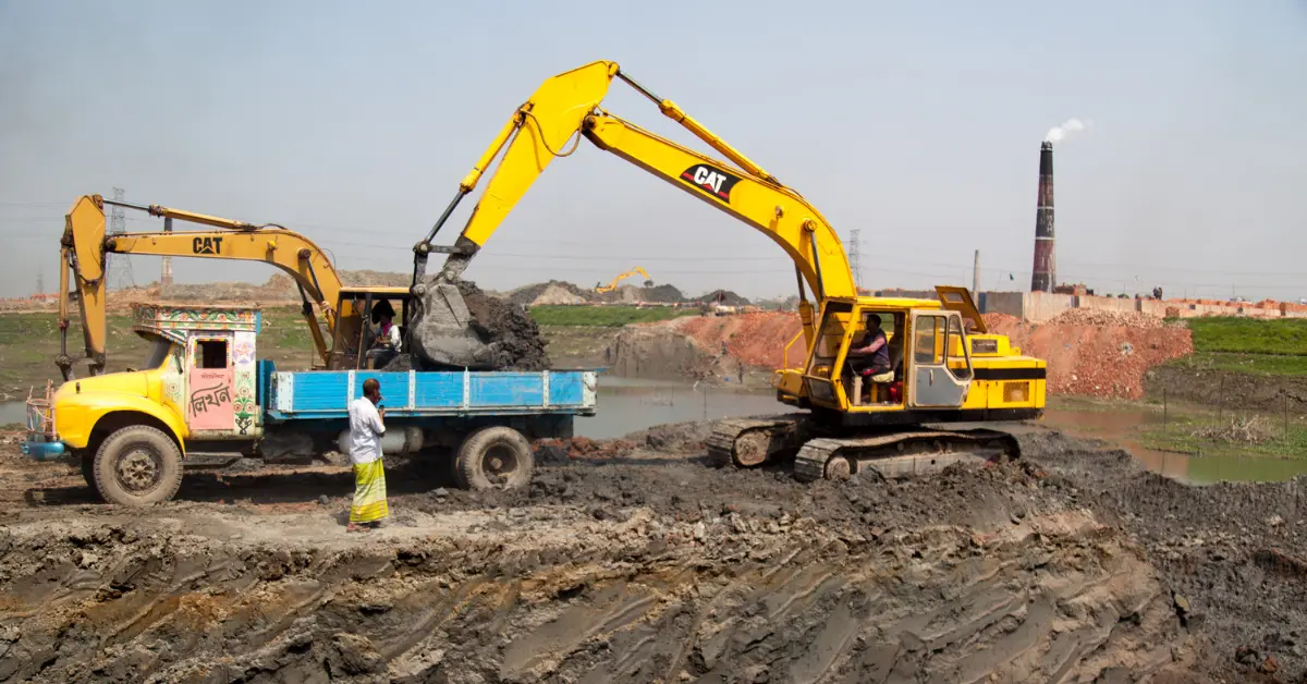 Clean fill dirt loaded into a dump truck by an excavator at a muddy construction site near a brick factory.