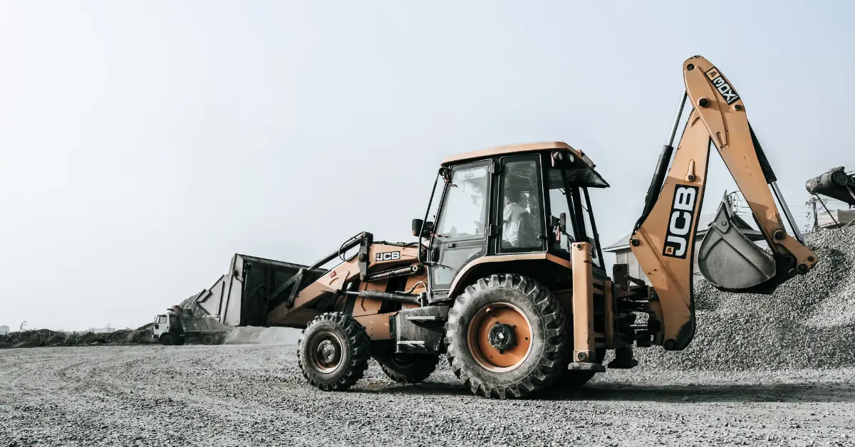 Clean fill dirt delivery using a backhoe loader at a gravel worksite with trucks in the background.