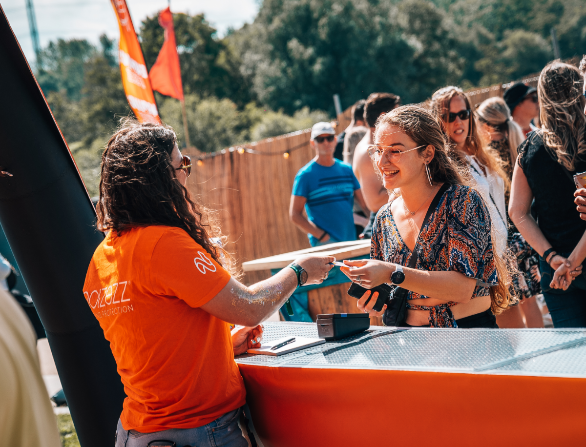 Een meisje geeft aan een ander meisje over de balie een zakje earbeanz op een festival.