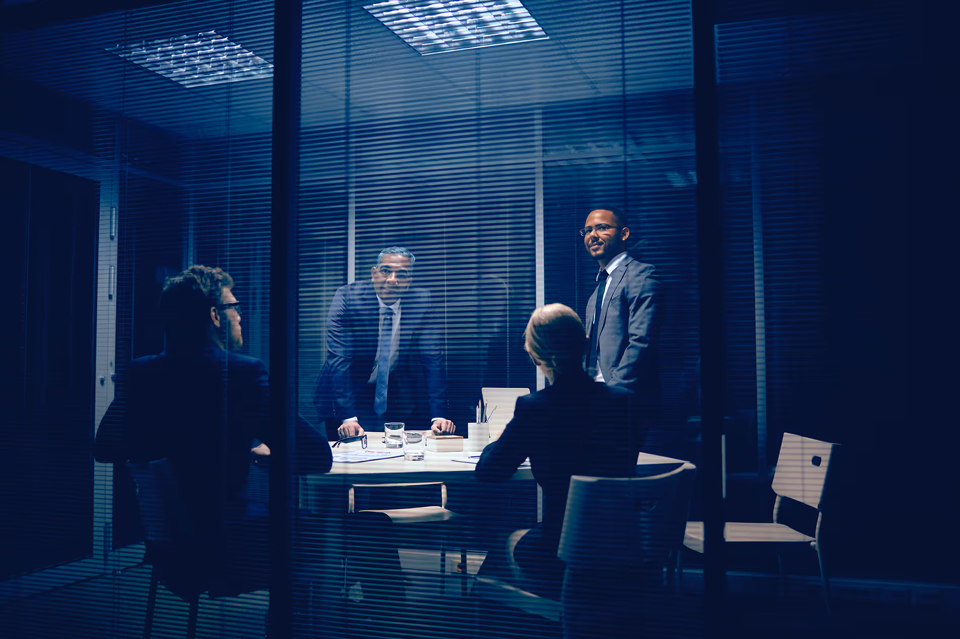 Four business professionals in suits having a meeting in a glass-walled conference room at night.