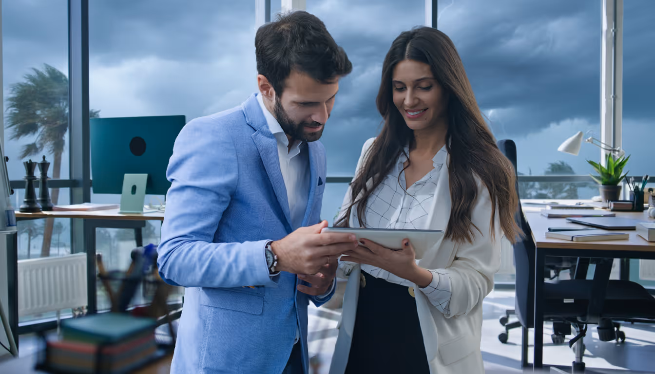 Two business professionals, a man in a blue blazer and a woman in a white blazer, smiling and looking at a tablet in a modern office with stormy weather outside.