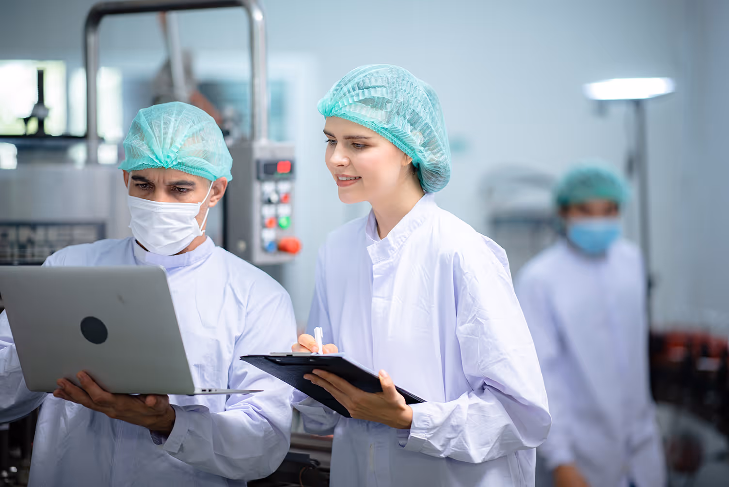 Two food technicians in protective clothing and hairnets inspecting production data on a laptop and clipboard in a factory.