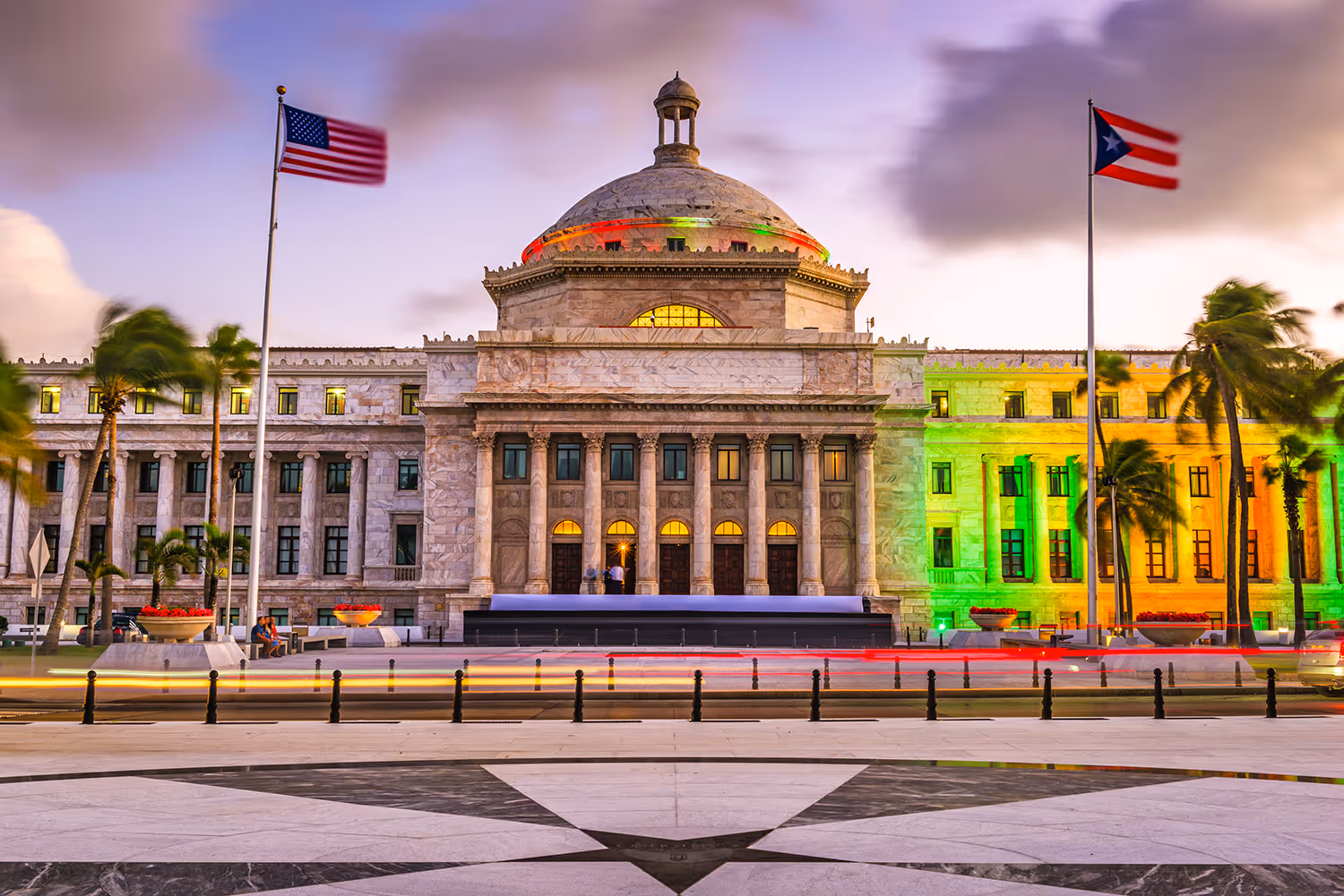 The Puerto Rico Capitol building illuminated with green and yellow lights, flanked by the US and Puerto Rican flags at dusk.
