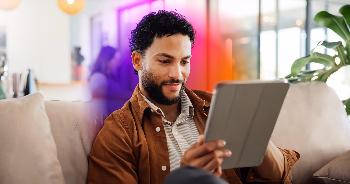 Man with curly black hair and beard sitting on a beige couch, wearing a brown jacket over a striped shirt, looking at a tablet in his hands, with a colorful blurred background and green plant on the right.