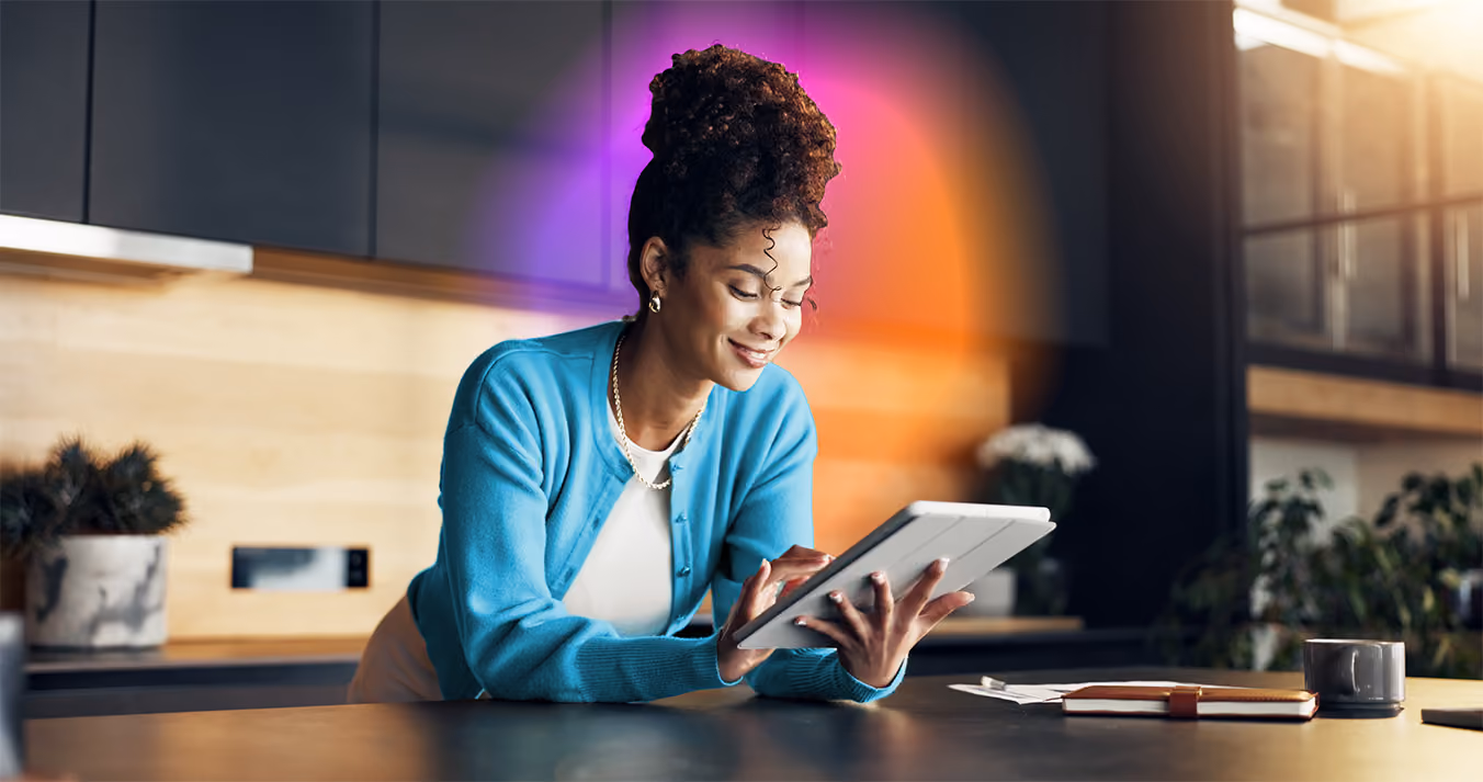 Woman with curly hair in a bun wearing a blue cardigan leaning on a kitchen counter, smiling while using a tablet, with a notebook, pen, and mug nearby.