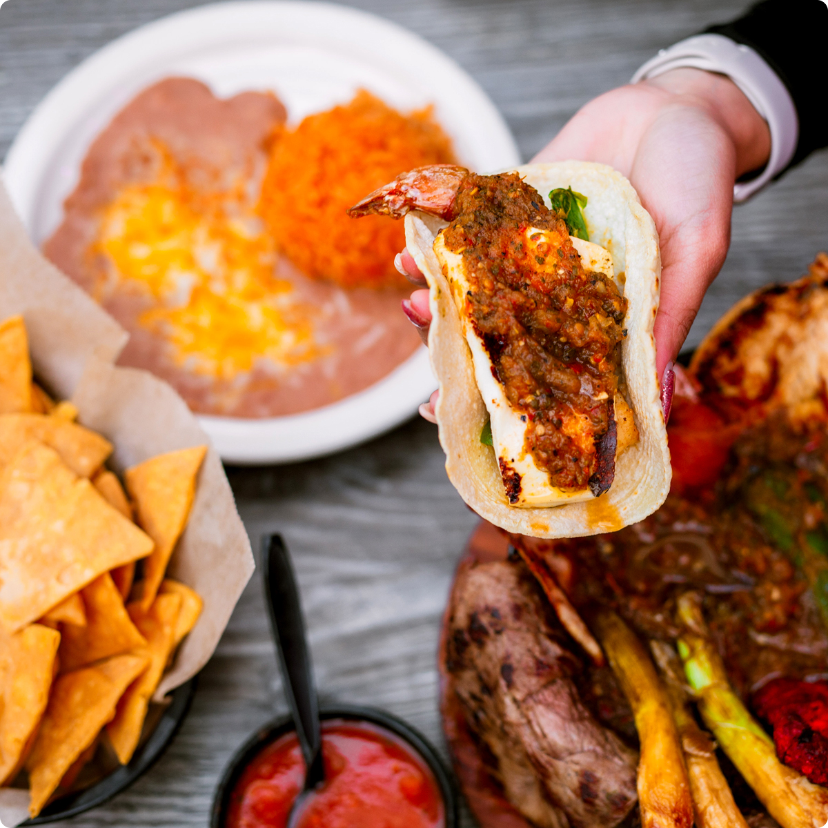 Shrimp tacos with queso being offered. Chips, beans and chile are in the background.