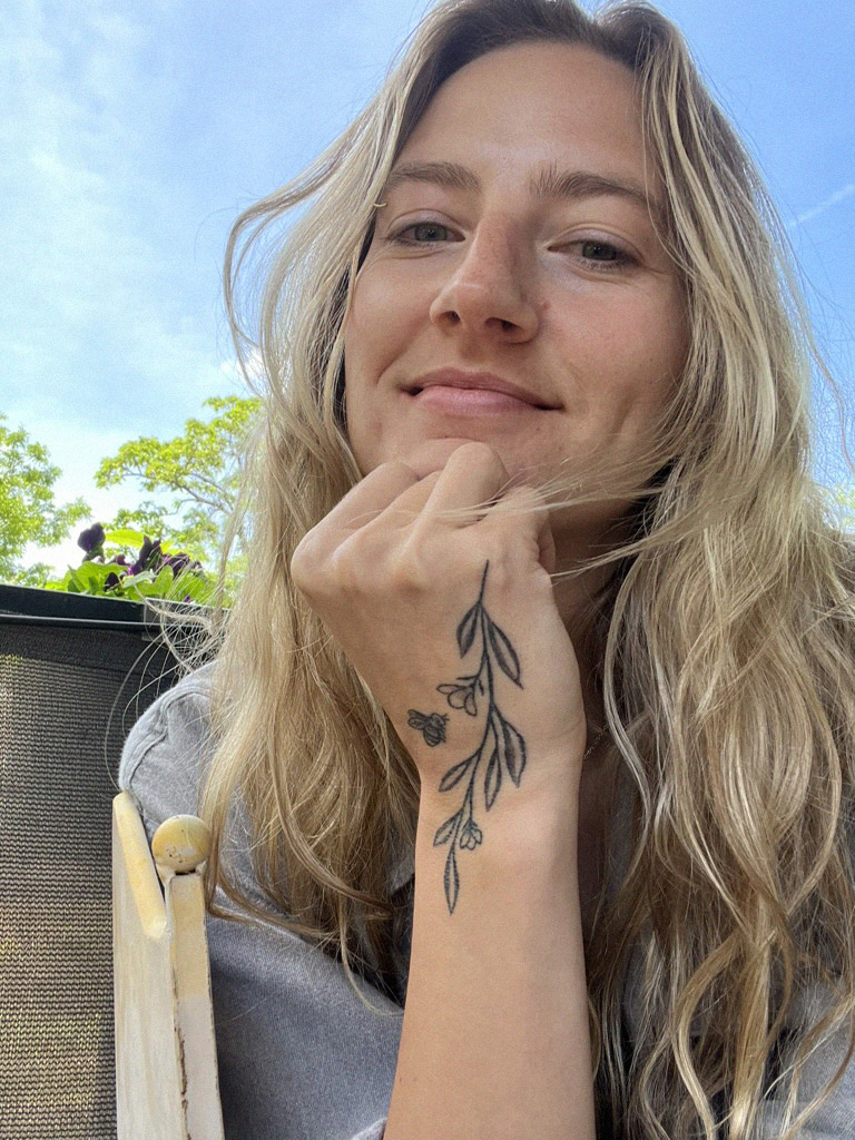 Smiling woman with long blonde hair resting her chin on her hand, showing a tattoo of a floral branch and a bee on her wrist, outdoors with trees in the background.