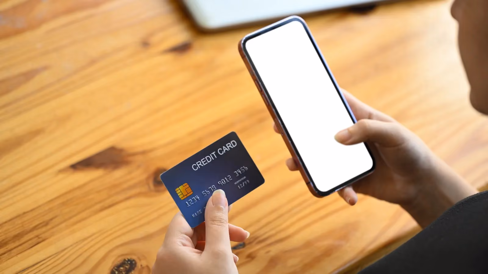 Tangan memegang kartu kredit dan ponsel dengan layar kosong di atas meja kayu, ilustrasi proses pembayaran digital dan fitur penyimpanan kartu/tokenisasi.