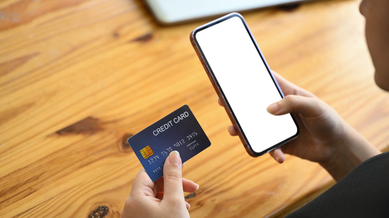 Tangan memegang kartu kredit dan ponsel dengan layar kosong di atas meja kayu, ilustrasi proses pembayaran digital dan fitur penyimpanan kartu/tokenisasi.