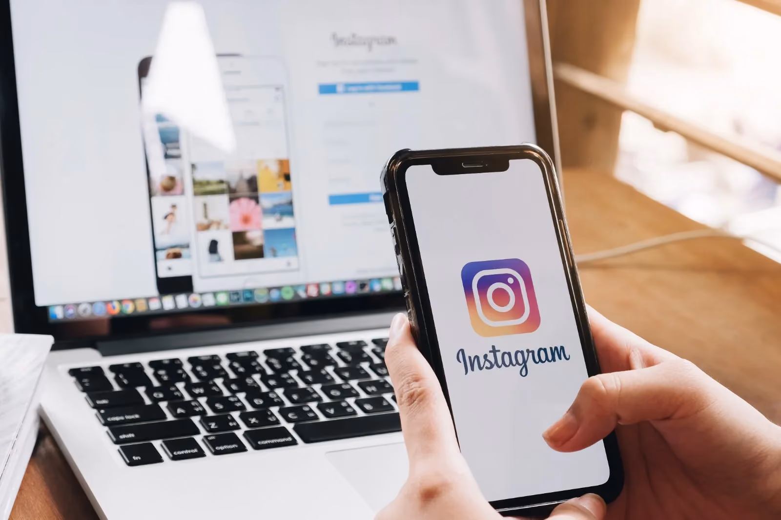 Seorang pengguna memegang smartphone dengan aplikasi Instagram yang ditampilkan di layar, sementara di latar belakang terlihat halaman Instagram di laptop, menunjukkan berbagai foto dan tombol login.
