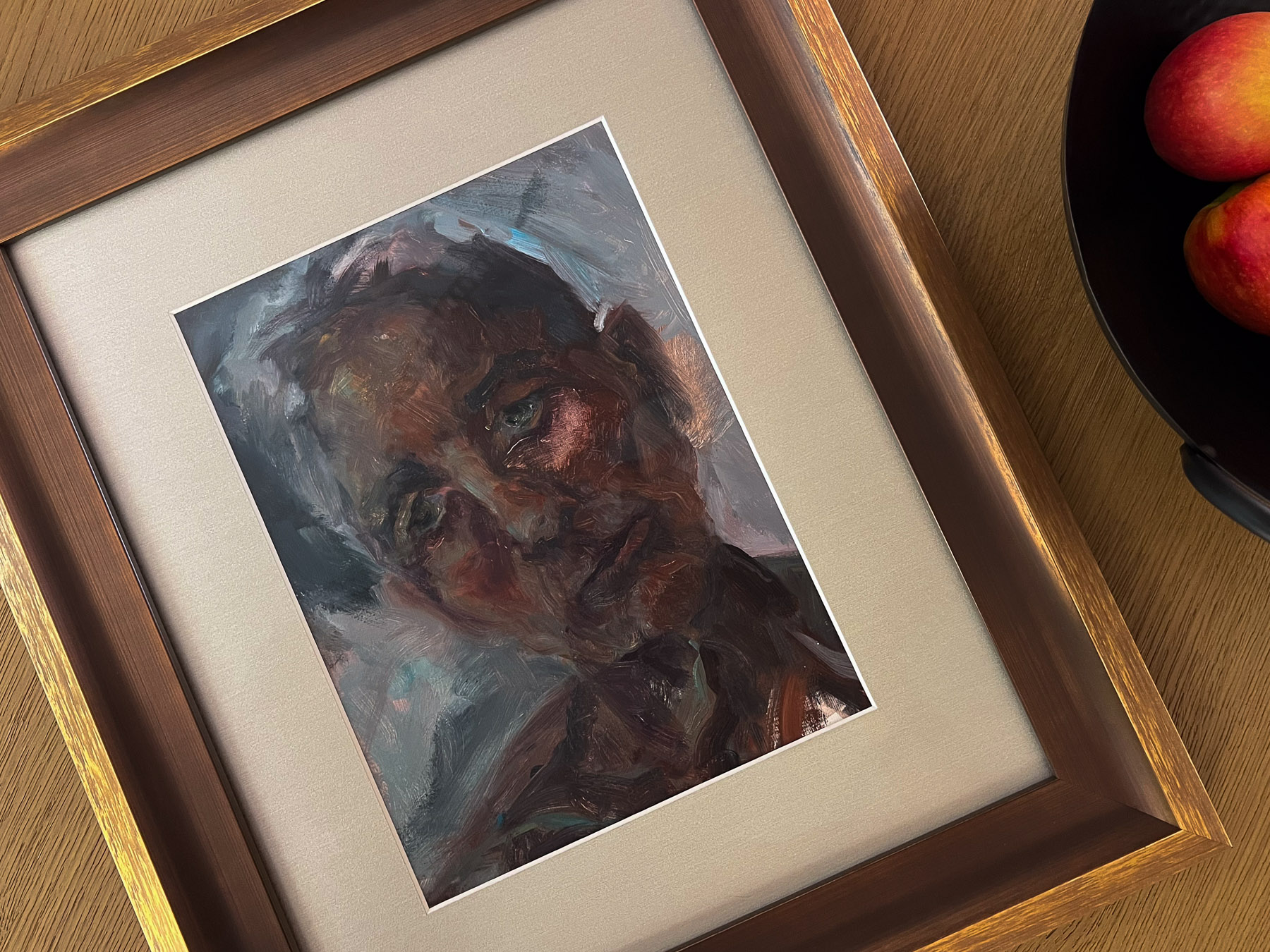 A framed, portrait-oriented painting of a person with dark, expressive brushstrokes sits on a wooden table, next to a black bowl holding two apples.