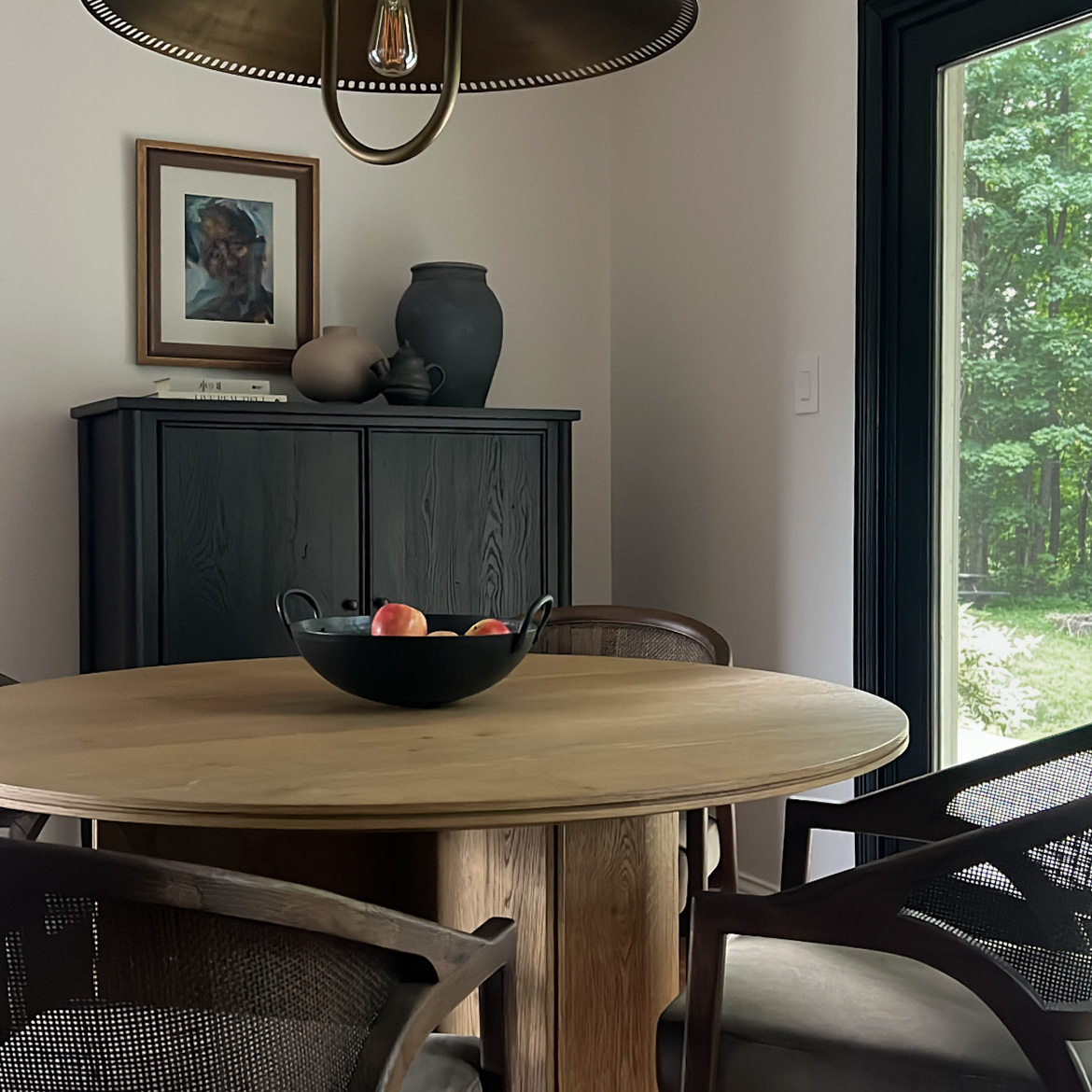 Round wooden dining table with a black bowl of fruit and chairs beside a cabinet and window.
