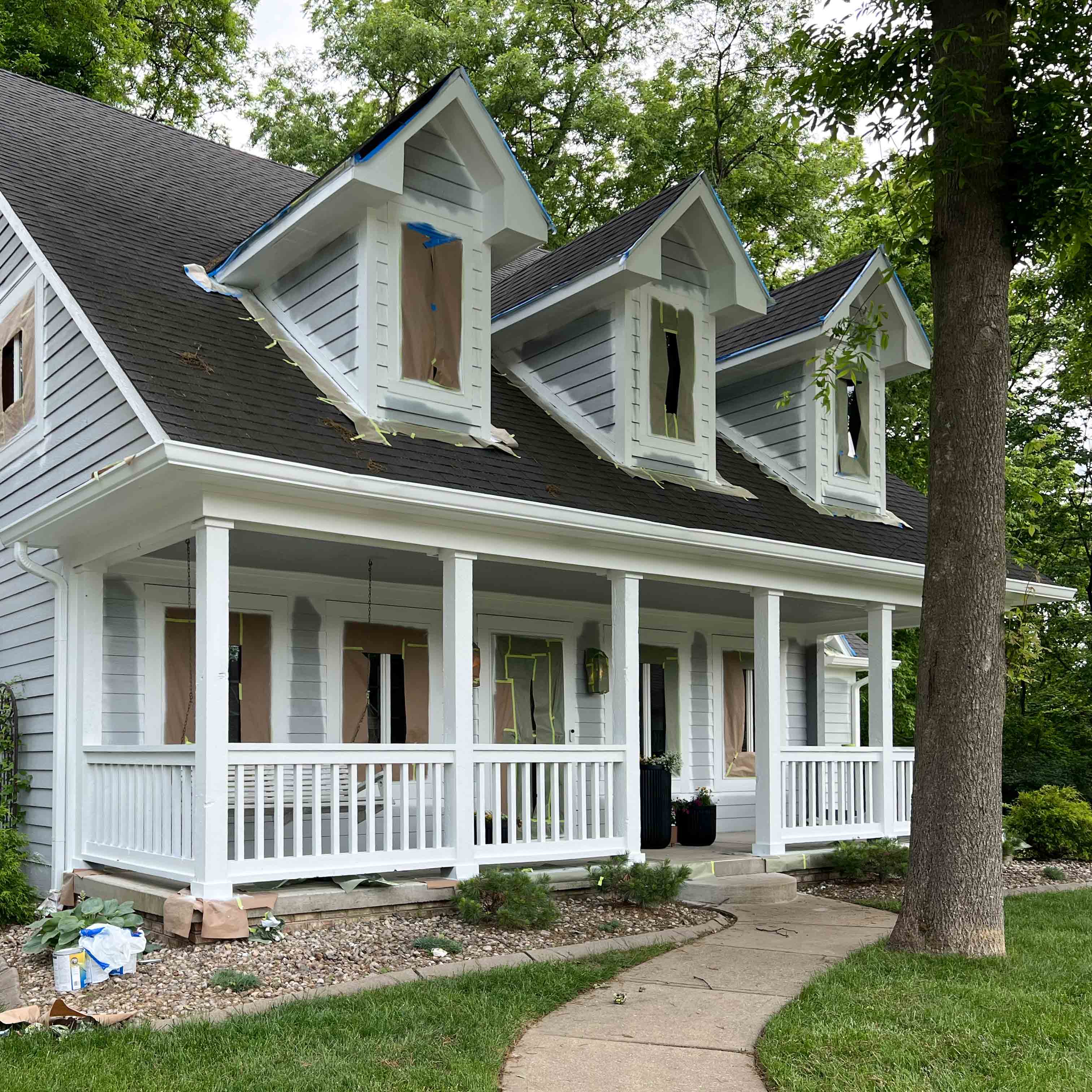 White house with light blue siding and covered porch undergoing exterior painting with windows taped off.