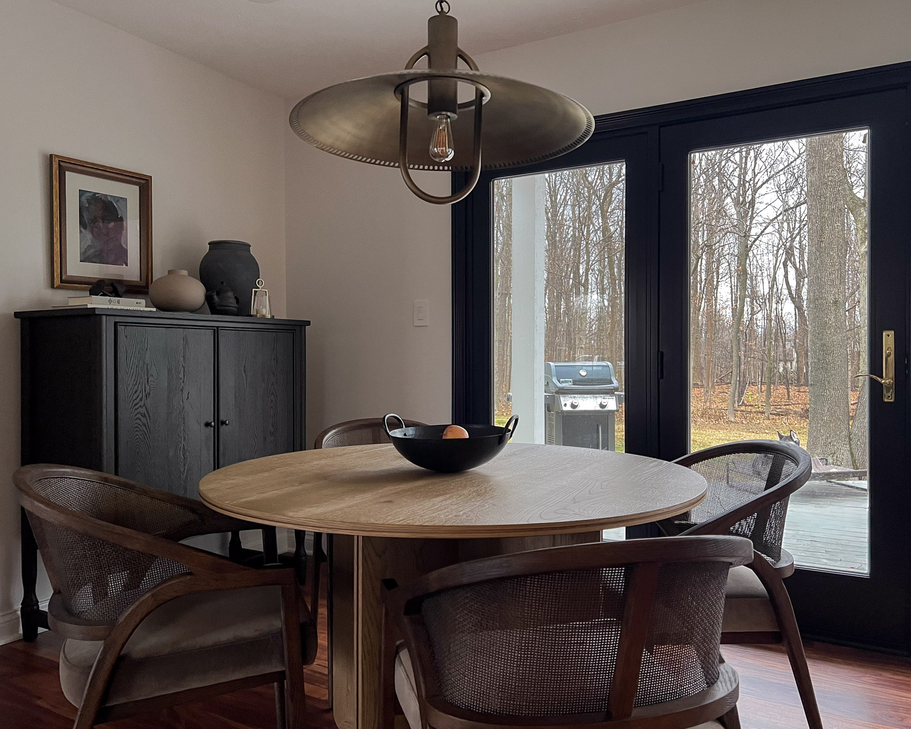 Round oak dining table with cane-back chairs and a pendant light, next to a dark cabinet with vases and framed art, beside French doors overlooking a wooded backyard.