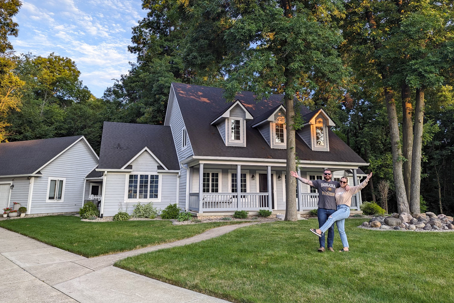 A couple stands on the front lawn with arms raised, celebrating in front of a white Cape Cod-style house with a front porch, dormer windows, and dark roof, surrounded by tall trees at dusk.