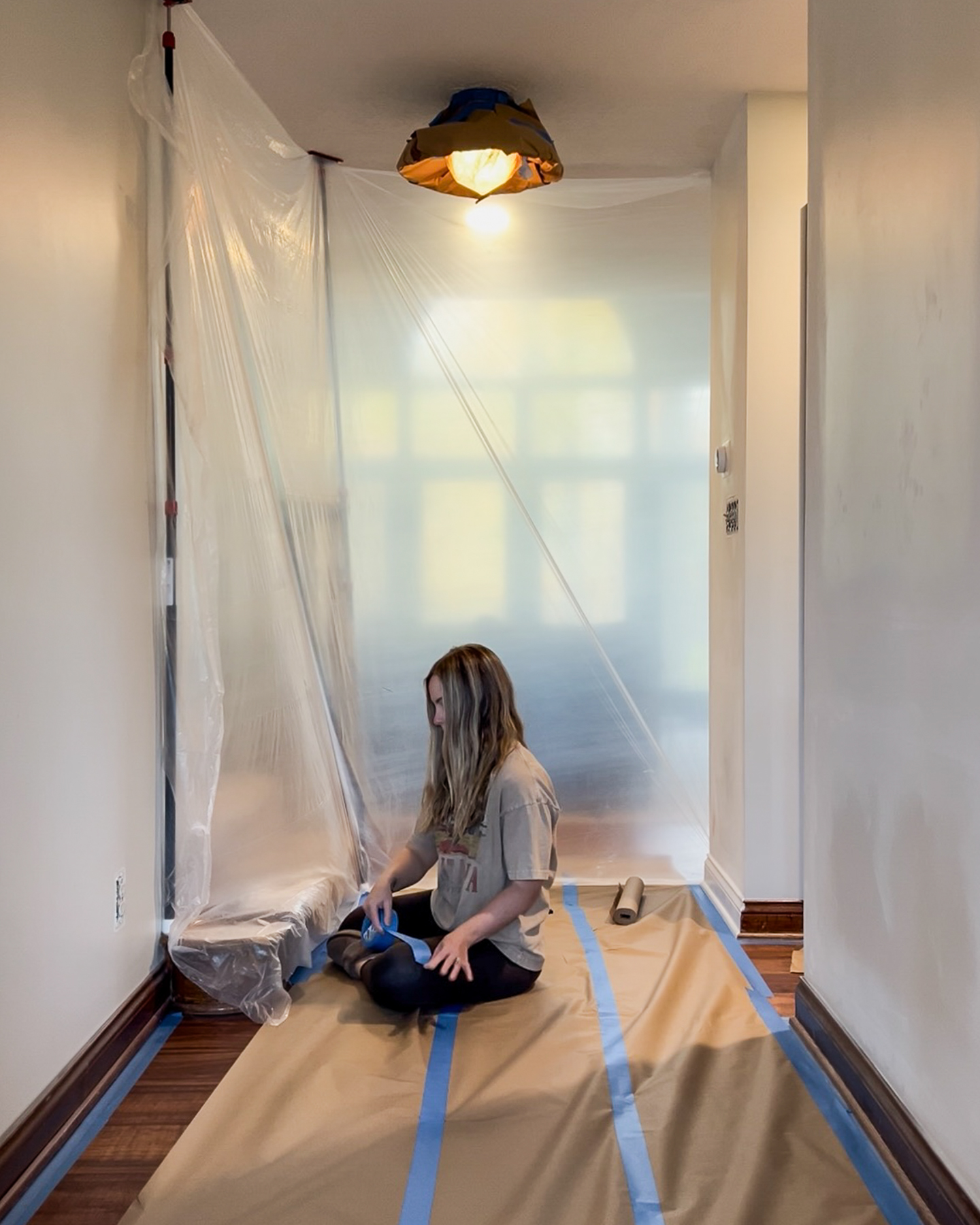 A woman kneels on a drop cloth in a hallway, painting trim, with plastic sheeting taped up and blue painter's tape lining the floor edges.