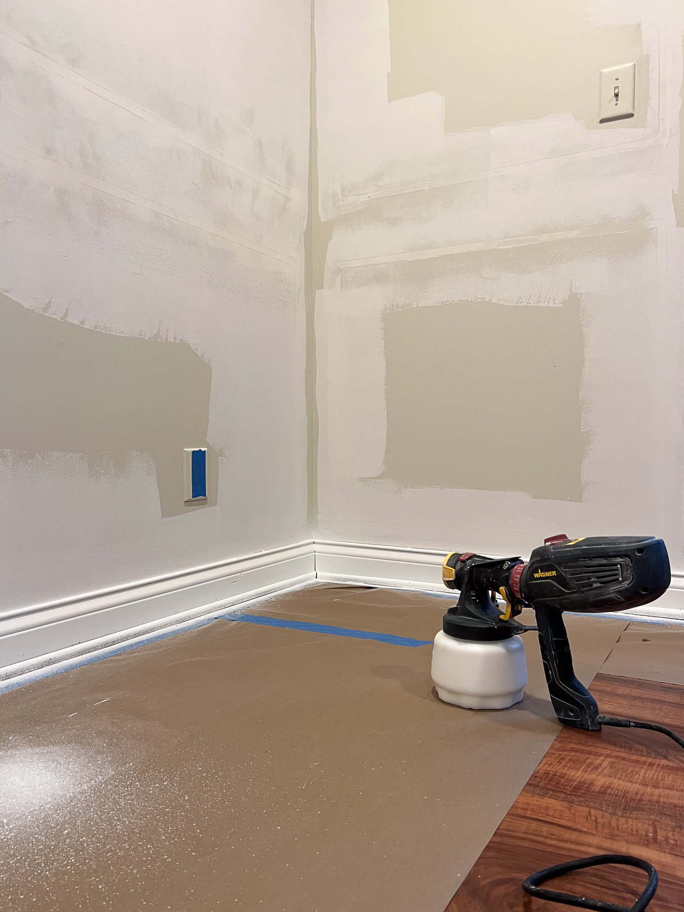 A paint sprayer sits on a floor protected by paper covering, in a room with test paint patches on the walls and an outlet cover removed.