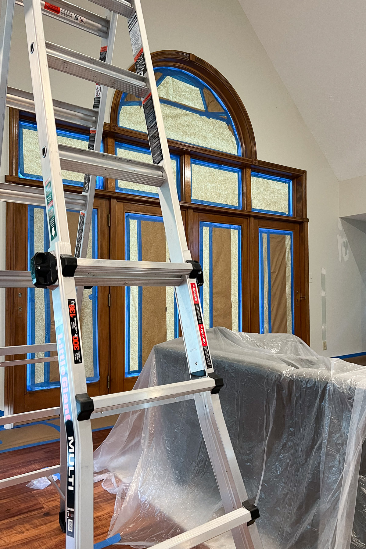 An aluminum ladder stands in a living room with an arched window and wood-paneled doors, all edges taped off in blue and nearby furniture covered in plastic sheeting.