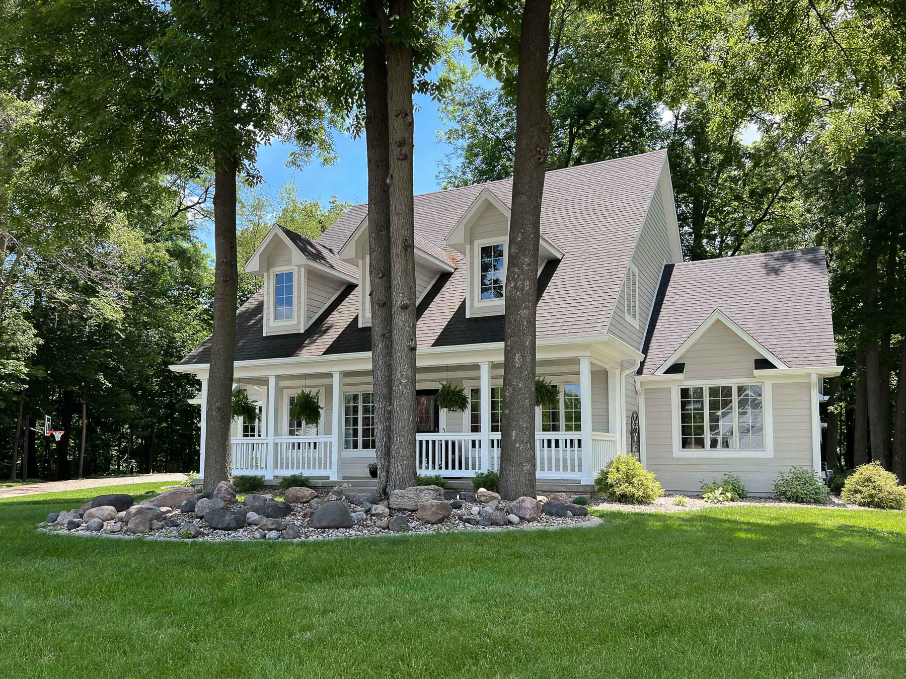 A beige Cape Cod-style home with a wraparound porch, dark shingle roof, and dormer windows, framed by tall pine trees and a landscaped rock garden on a green lawn.