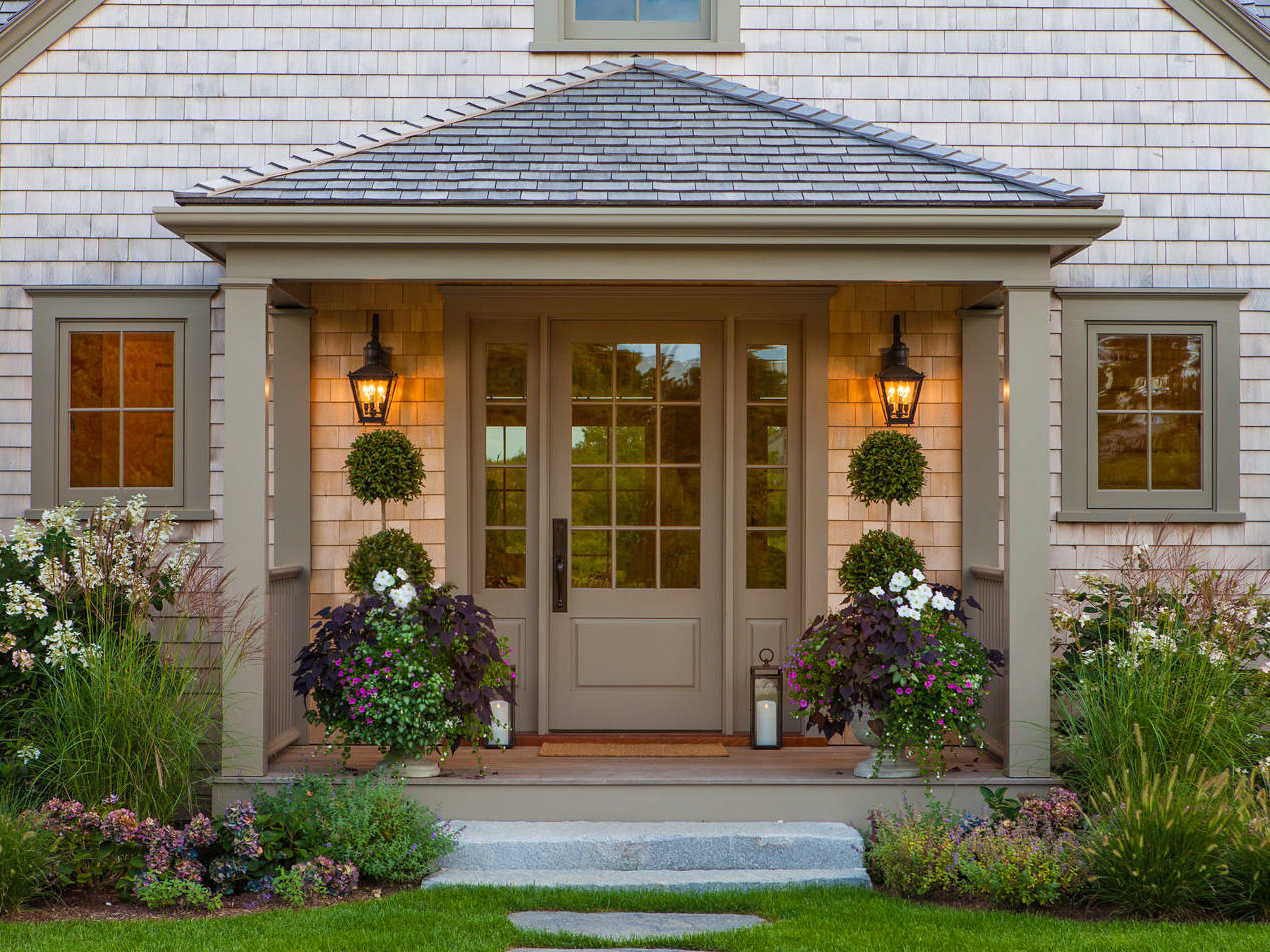 A Craftsman or Coastal style home entryway with cedar shingle siding and dark sage trim. The glass-panel front door has matching sidelights and is framed by a covered porch with two glowing lantern sconces, potted topiaries, vibrant flower planters, and stone steps leading up from a green lawn.