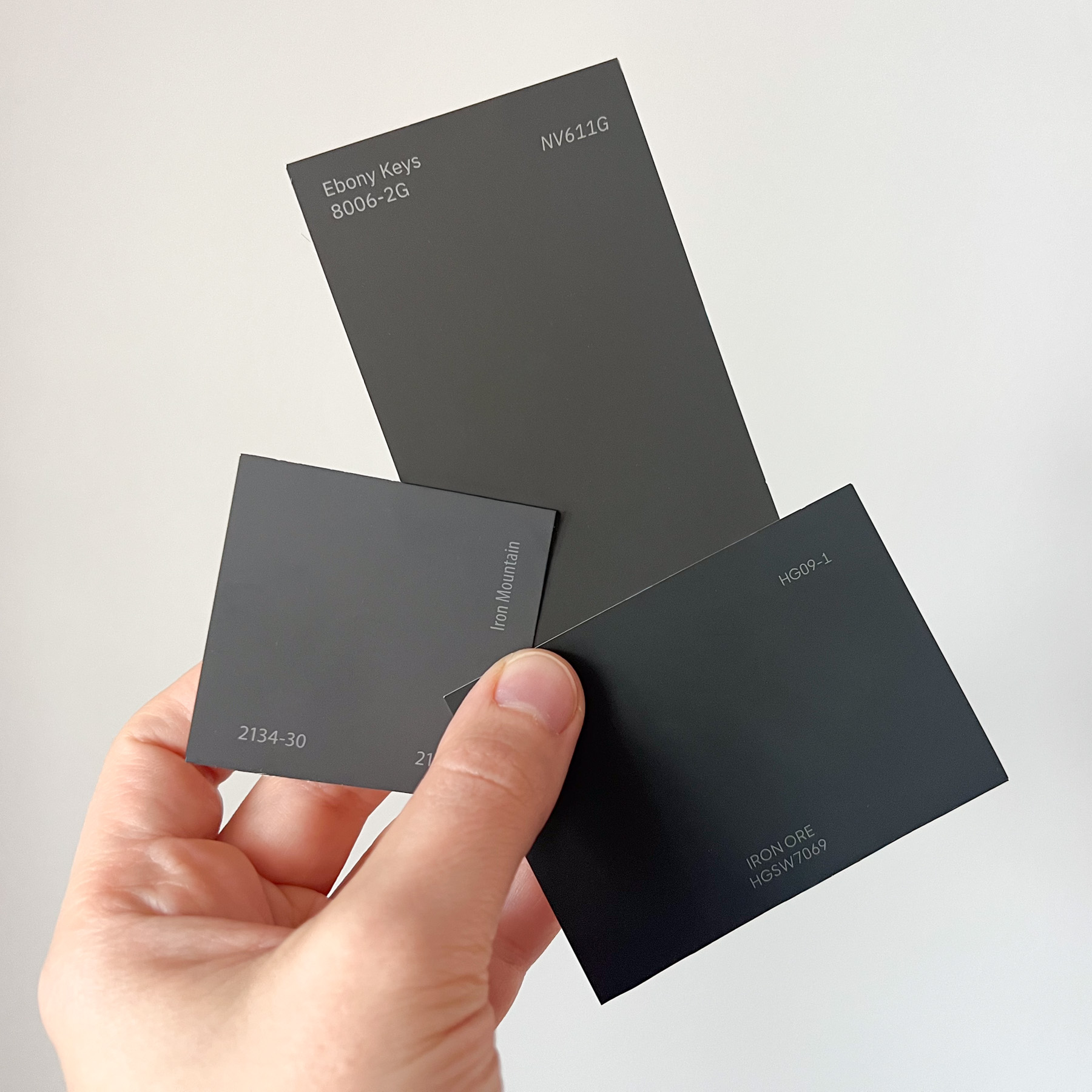 A hand holding three dark paint color swatches against a plain white background. The swatches are various shades of charcoal and near-black. The middle swatch is titled "Ebony King," the left is "Iron Mountain," and the right is labeled "Iron Ore."
