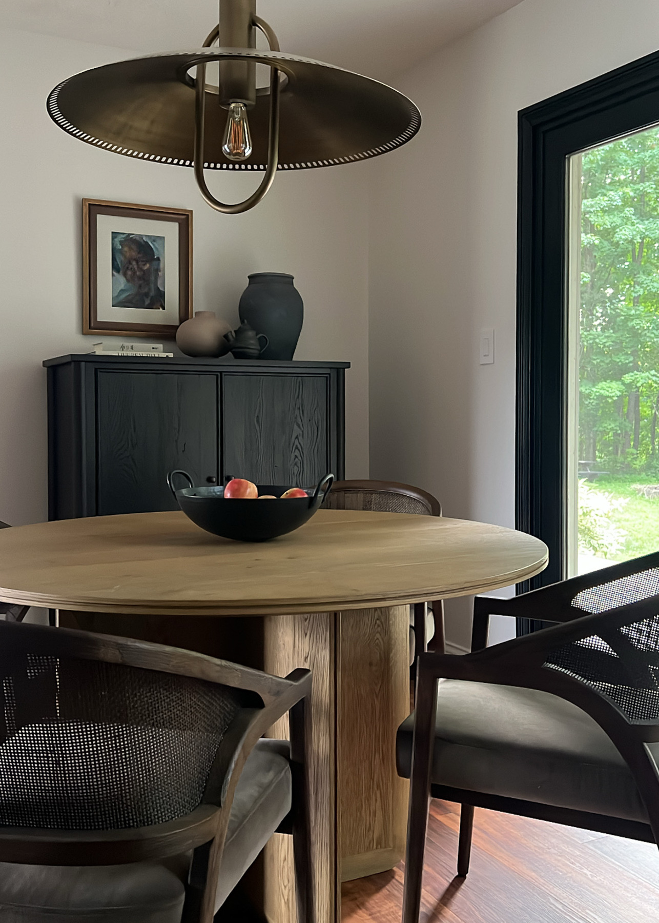 A dining room featuring a round wooden table with a bowl of apples, surrounded by dark cane-backed chairs. A black bar cabinet with vases and a framed portrait is against the left wall.