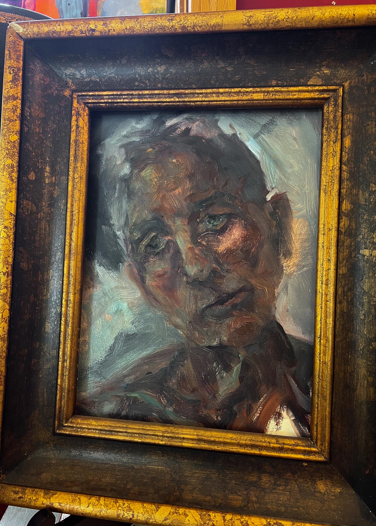 A close-up of a framed painting of a solemn-looking person with dark, expressive brushstrokes in a dark and distressed gold frame.