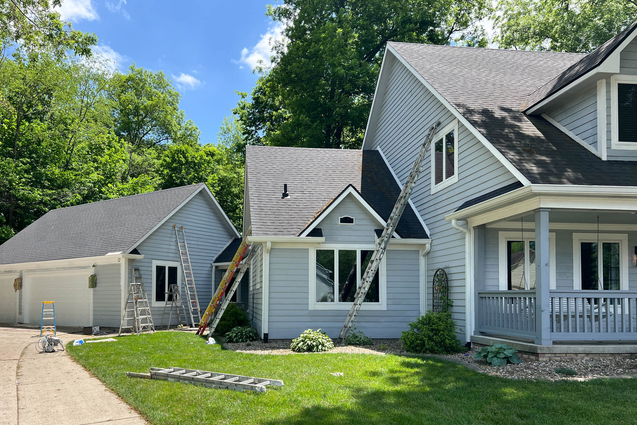 A two-story house with a blue exterior and black roof. The house has a porch and a garage attached to the side. There are trees in the background.