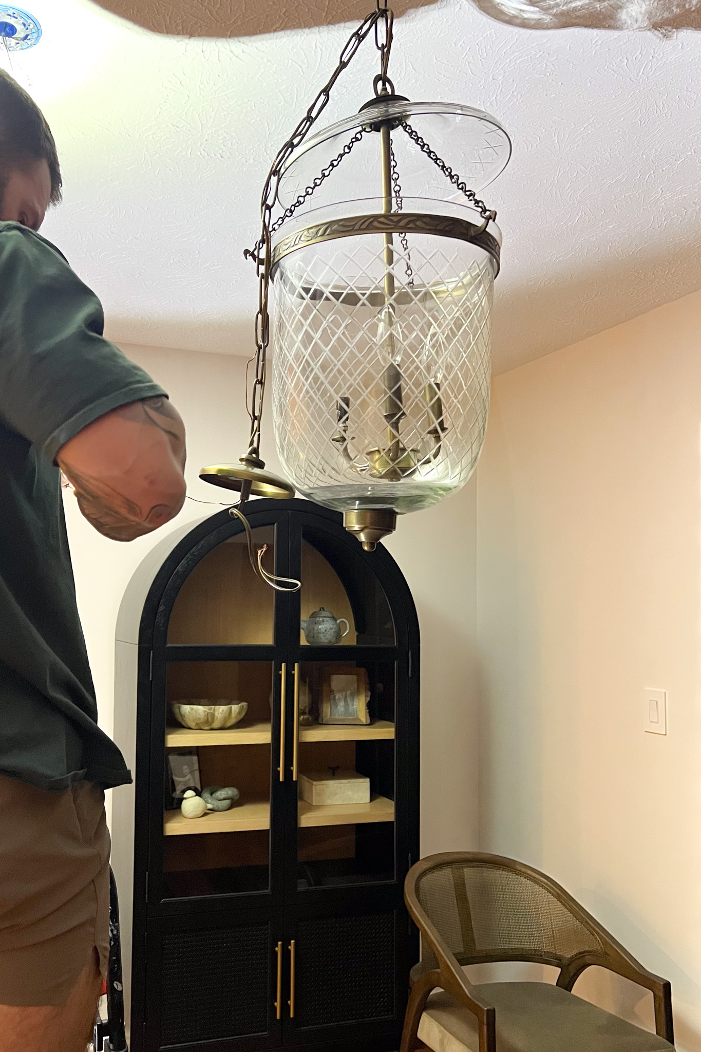 A person installs a large, antique brass and etched glass lantern pendant light over a black arched display cabinet and a cane-back chair.
