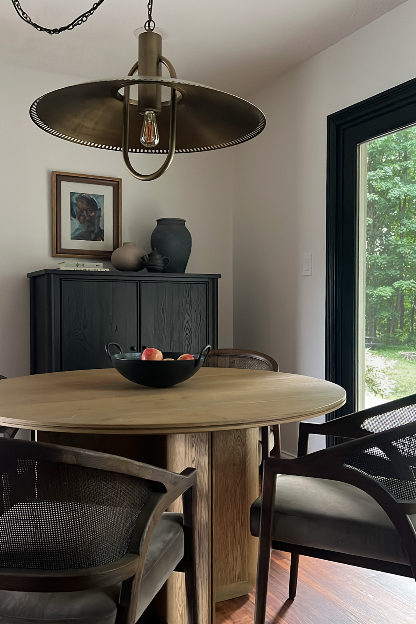 Overhead shot of a modern dining space with a brass pendant light, a black cabinet holding decor, and a wooden table with a bowl of fruit.