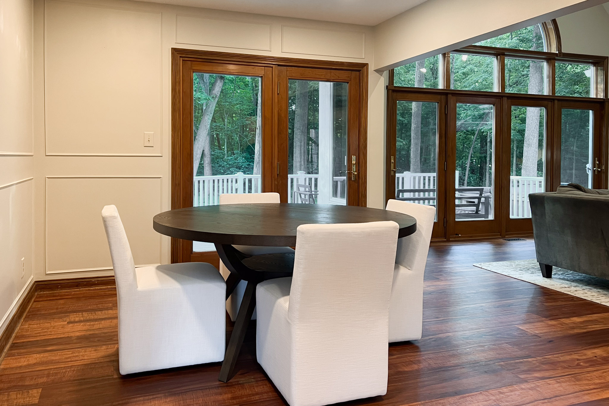 Dining room with a dark round table and four white upholstered chairs, wood paneled doors leading to a deck, and hardwood floors.