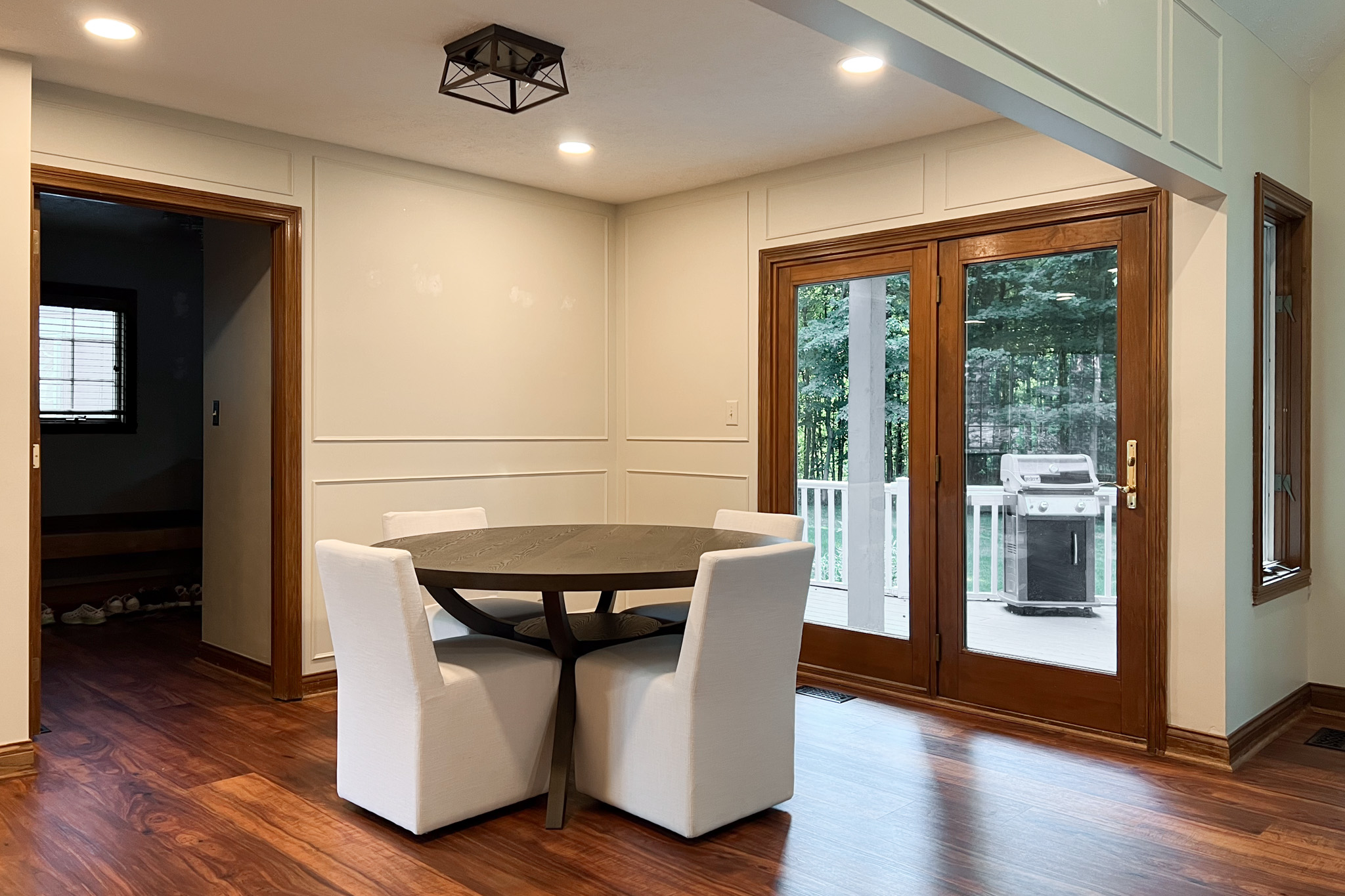 Interior view of a dining area with dark wood floors, white paneled walls, and a round dark table with four white upholstered chairs under a geometric light fixture.