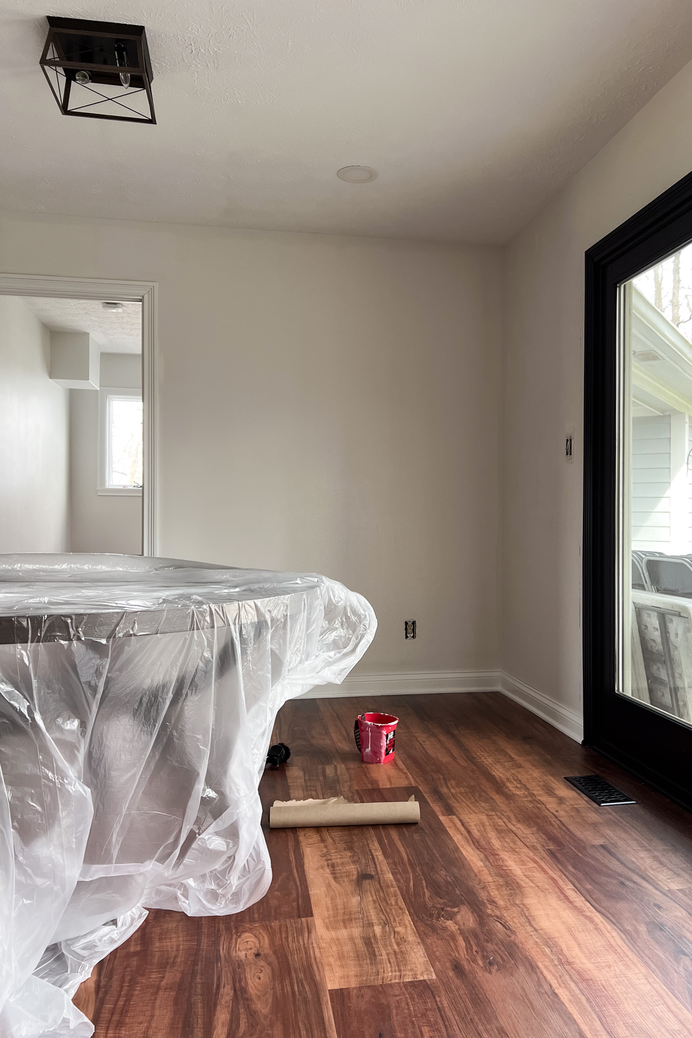 Interior view of a dining nook in renovation; a table is covered in plastic, showing dark wood floors, light walls, and a black-trimmed door.