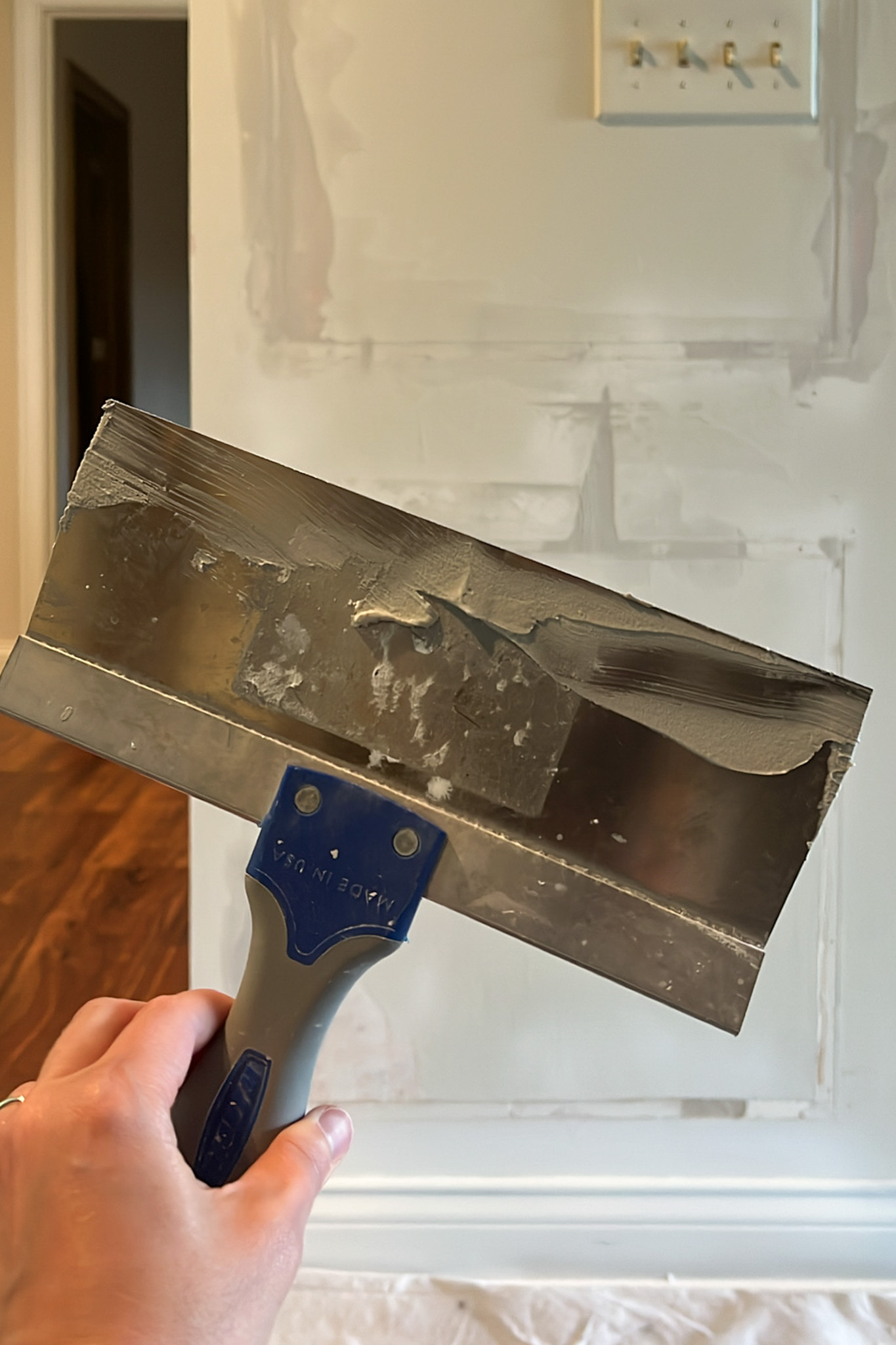 A hand holds a drywall knife with joint compound, used for patching a wall.
